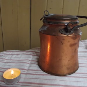 May include: A copper milk can with a hinged lid and a handle. The can has a worn, aged patina and is sitting on a striped tablecloth.
