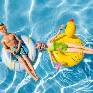 May include: Two dolls floating in a pool. One doll is on a blue and white inflatable ring, wearing sunglasses and swim trunks. The other doll is on a yellow duck-shaped float, wearing a green swimsuit and headphones. The water is a bright blue.