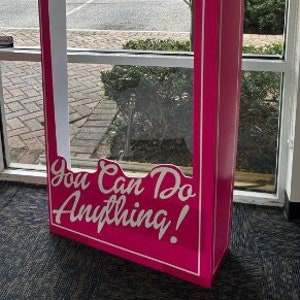 May include: A pink cardboard photo booth cutout with a white frame and the text "You Can Do Anything!" in white lettering.