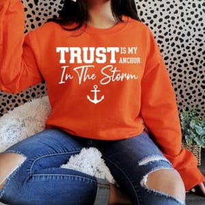 Peut inclure: Un sweat-shirt orange avec l'inscription blanche "TRUST is my anchor In The Storm" et un motif d'ancre blanc. Le sweat-shirt est porté avec un jean bleu.