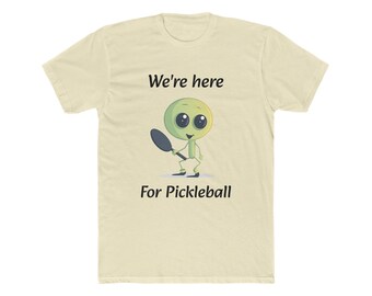 May include: A light beige t-shirt featuring a green alien holding a paddle and the text "We're here for Pickleball".