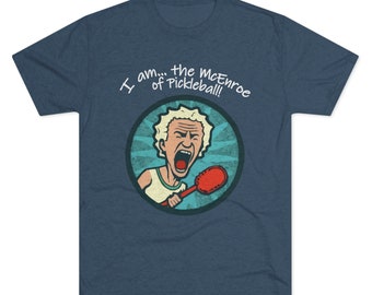 May include: A blue t-shirt featuring a cartoon image of a man with white hair shouting while holding a red pickleball paddle. The text "I am... the McEnroe of Pickleball!" is printed in white on the shirt.