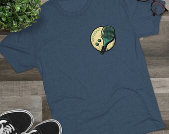 May include: A dark blue t-shirt featuring a vintage-style graphic of a pickleball paddle and balls. The graphic is in yellow, green, and brown colours. The text "Pickleball" is written in a circular design around the paddle and balls.