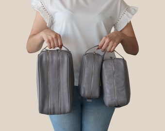 May include: Three gray leather travel bags of varying sizes are held up. Each bag has a silver zipper and a small handle. The bags are rectangular with rounded corners. The bags are a light gray color.