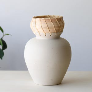 May include: A cream-colored ceramic vase with a woven, light brown rim. The vase has a rounded body and a textured surface. The woven rim adds a natural, rustic element to the design. The vase is displayed on a white surface.