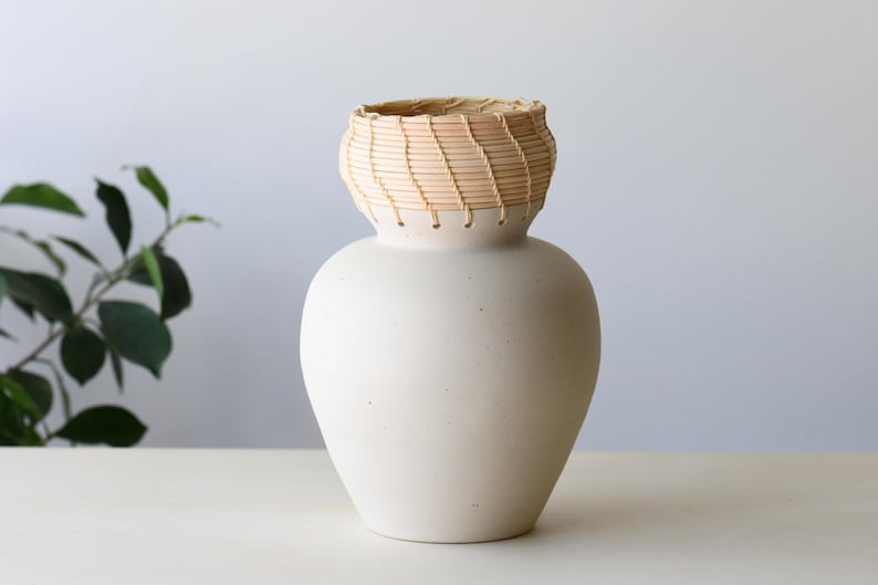 May include: A cream-colored ceramic vase with a woven, light brown rim. The vase has a rounded body and a textured surface. The woven rim adds a natural, rustic element to the design. The vase is displayed on a white surface.