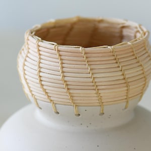 May include: A small, handcrafted basket with a woven, light-colored reed design. The basket sits atop a white ceramic base. The basket's rim and the base's connection point are visible in this close-up shot.