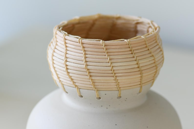 May include: A small, handcrafted basket with a woven, light-colored reed design. The basket sits atop a white ceramic base. The basket's rim and the base's connection point are visible in this close-up shot.