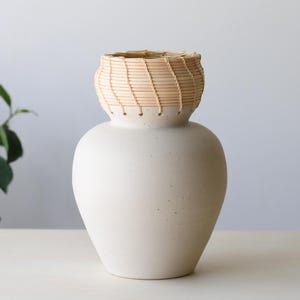 May include: A cream-colored ceramic vase with a woven, light brown basket-like top. The vase has a rounded body and a textured surface. The top is a lighter color than the body. The vase is sitting on a white surface.