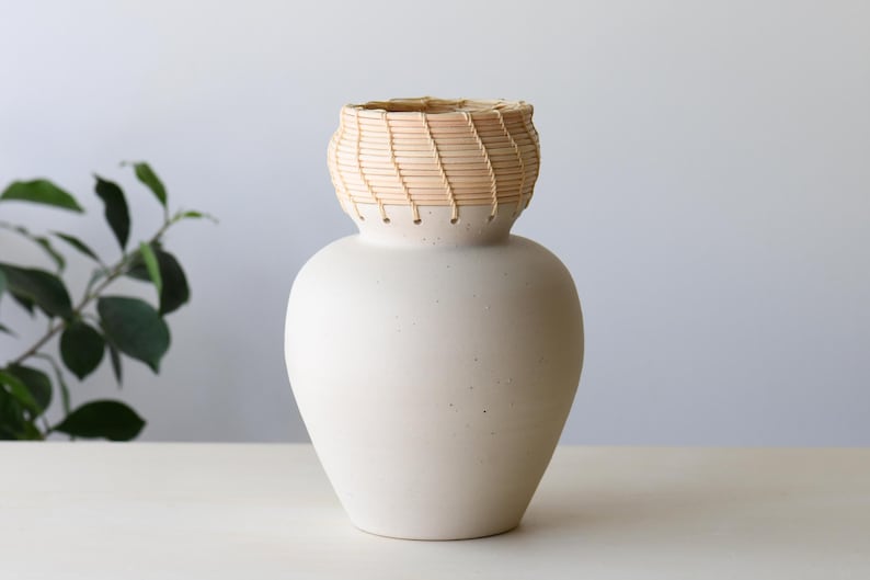 May include: A cream-colored ceramic vase with a woven, light brown basket-like top. The vase has a rounded body and a textured surface. The top is a lighter color than the body. The vase is sitting on a white surface.