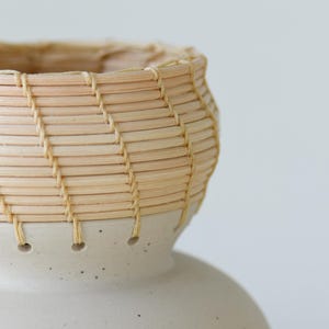 May include: A handcrafted ceramic vase with a woven basket-like top. The vase features a light beige color with a speckled base and a woven top made of thin, light-colored reeds. The design is simple and elegant, suitable for home decor.