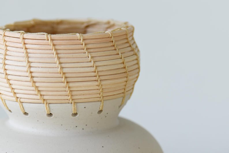 May include: A handcrafted ceramic vase with a woven basket-like top. The vase features a light beige color with a speckled base and a woven top made of thin, light-colored reeds. The design is simple and elegant, suitable for home decor.