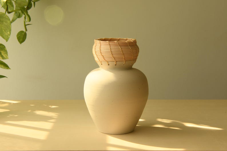 May include: A cream-colored ceramic vase with a woven, light brown top. The vase has a rounded body and sits on a light-colored wooden surface. The background is a soft, muted green. The vase is a decorative home decor item.