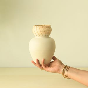 May include: A cream-colored ceramic vase with a woven, light brown top section. The vase is held by a person's hand, adorned with gold bracelets. The background is a soft, neutral color, with a hint of greenery on the left.