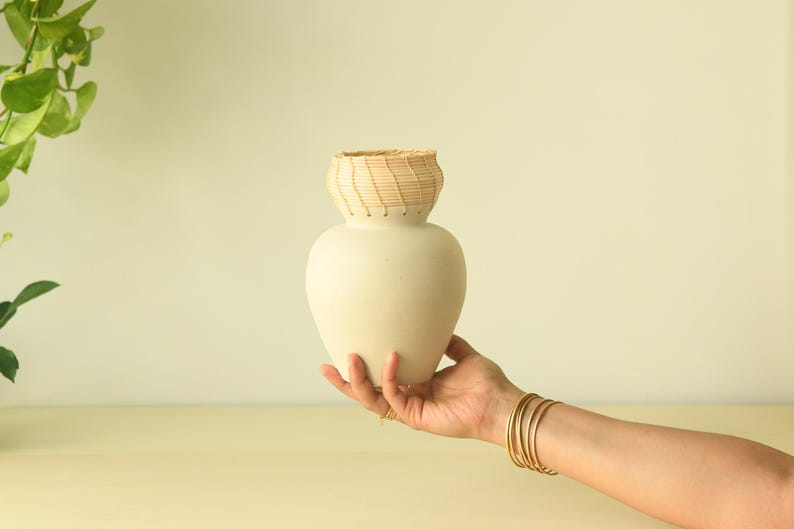 May include: A cream-colored ceramic vase with a woven, light brown top section. The vase is held by a person's hand, adorned with gold bracelets. The background is a soft, neutral color, with a hint of greenery on the left.