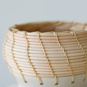 May include: A handcrafted, small woven basket with a light beige color. The basket is made of thin, natural-colored reeds and features a cream-colored ceramic base. The woven pattern creates a textured surface.