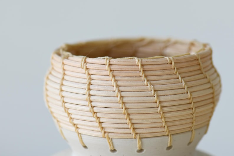 May include: A handcrafted, small woven basket with a light beige color. The basket is made of thin, natural-colored reeds and features a cream-colored ceramic base. The woven pattern creates a textured surface.