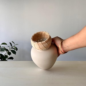 May include: A cream-colored, heart-shaped ceramic vase topped with a small, woven basket. The basket is made of thin, light-colored wooden strips. A gold bracelet adorns the wrist holding the vase. The objects are on a light-colored surface.