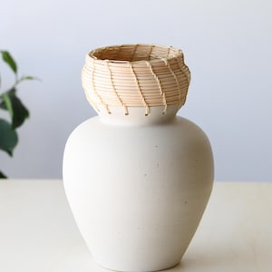 May include: A white ceramic vase with a woven, light brown basket-like top. The vase has a rounded body and a slightly flared opening. The woven top adds a natural texture to the smooth, matte finish of the vase. A decorative piece for the home.