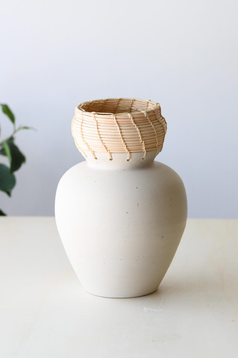 May include: A white ceramic vase with a woven, light brown basket-like top. The vase has a rounded body and a slightly flared opening. The woven top adds a natural texture to the smooth, matte finish of the vase. A decorative piece for the home.