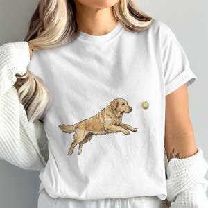 Könnte beinhalten: Ein weißes Kurzarm-T-Shirt mit einer grafischen Illustration eines goldfarbenen Hundes, der durch die Luft auf einen kleinen gelben Tennisball zuspringt. Der Hund ist in einer Sprungbewegung mit ausgestrecktem Körper und leicht erhobenem Schwanz dargestellt.