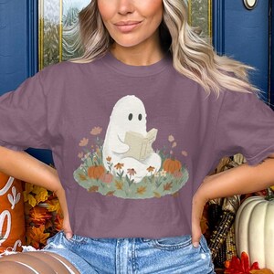 Può includere: Una t-shirt color malva a maniche corte con una stampa grafica raffigurante un fantasma bianco seduto in un campo di fiori autunnali e zucche mentre legge un libro aperto. Il disegno è centrato sul petto del capo, dotato di girocollo.