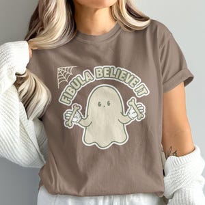 May include: A brown t-shirt featuring a graphic of a light-colored ghost holding two bones. The text "FIBULA BELIEVE IT" is arched above the ghost in a light-colored font, accompanied by a small spiderweb illustration in the upper left corner.