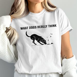 Könnte beinhalten: Ein weißes T-Shirt mit kurzen Ärmeln und einer schwarzen Grafik eines Hundes, der im Boden gräbt. Über dem Hund steht der Text WHAT DOGS REALLY THINK in einer fetten, schwarzen, serifenlosen Schrift. Das Shirt hat einen Rundhalsausschnitt.