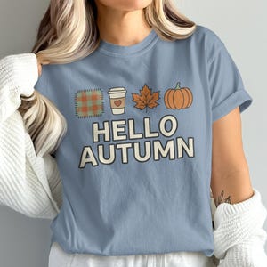 Könnte beinhalten: Ein staubblaues Kurzarm-T-Shirt mit einem Grafikdruck, der ein kariertes Quadrat, einen Kaffeebecher mit Herz, ein Ahornblatt und einen orangefarbenen Kürbis zeigt. Unter den Symbolen steht der Text HELLO AUTUMN in cremefarbener Schrift.