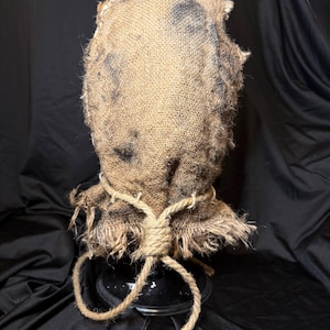 Op de afbeelding: Een beige jute zakmasker met een ruw, getextureerd oppervlak en donkere, onregelmatige vlekken. De onderkant van het masker is samengebonden met een dik, geknoopt touw van jute dat in twee losse, rafelige uiteinden naar beneden hangt.
