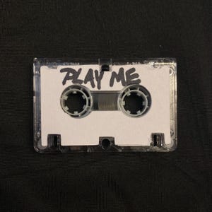 May include: A clear cassette tape with a white label. The label has the words "PLAY ME" in black, handwritten-style lettering. The tape's spools are gray, and the case is transparent.