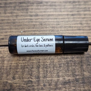Puede incluir: Un frasco de rodillo de vidrio marrón etiquetado como "Under-Eye Serum" para ojeras, líneas finas e hinchazón. El frasco tiene una tapa negra y está sobre una superficie de madera.