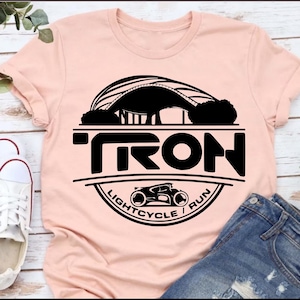 May include: A pink t-shirt with a black graphic design featuring the word "TRON" in a circle with a lightcycle and the text "Lightcycle / Run".