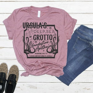 May include: A pink t-shirt with a black graphic design that says "Ursula's Deep Sea Grotto Clandestine Apothecary Bringing Aid to Poor Unfortunate Souls".