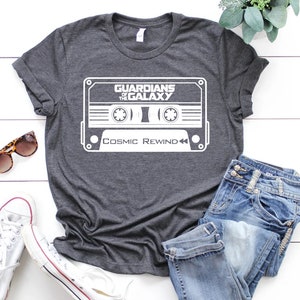 May include: A dark gray t-shirt with a white graphic of a cassette tape with the text "Guardians of the Galaxy" and "Cosmic Rewind" printed on it.