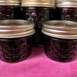May include: A collection of small glass jars filled with a dark, jam-like substance. The jars have silver lids and a diamond pattern on the glass. They are arranged on a pink surface, suggesting a homemade food product.