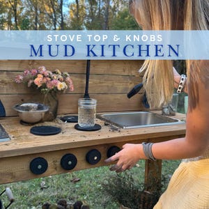 May include: A wooden mud kitchen with a stainless steel sink, black knobs, and a water tap. The text "STOVE TOP & KNOBS MUD KITCHEN" is displayed across the top. A person is shown interacting with the knobs.
