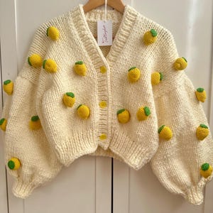 Op de afbeelding: Een crèmekleurige gebreide cardigan met een citroenmotief. De cardigan heeft een V-hals, lange mouwen en een knoopsluiting. De citroenen zijn gehaakt en hebben groene bladeren.