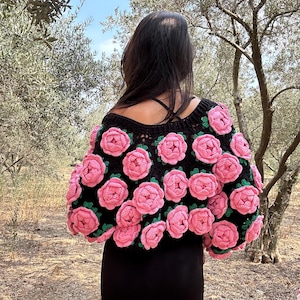 May include: Black crochet top adorned with pink rose appliques. The sleeves and neckline are trimmed with green. The top is worn over a black dress. The background features trees and a blue sky.