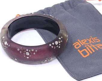 May include: A purple and clear Alexis Bittar bangle bracelet with embedded crystals. The bracelet is next to a gray pouch with the Alexis Bittar logo in orange. The bracelet has a dark interior.