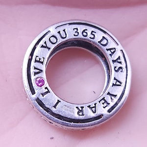 May include: Silver charm with the words "I love you 365 days a year" engraved around the edge. A small pink gemstone is set in the center of the charm.