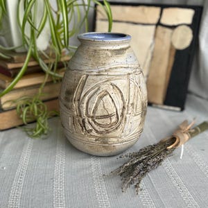 May include: A white ceramic vase with a blue rim and abstract brown markings. The vase is sitting on a white and gray striped surface.
