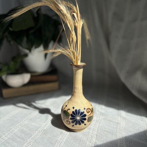 May include: A beige ceramic vase with a narrow neck and a round body. The vase is decorated with a blue flower and green and brown accents. It contains dried wheat stalks. The vase is sitting on a white surface.