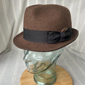 May include: A brown fedora hat with a black band and a small feather accent. The hat has a classic design and is displayed on a glass head form. The hat has the brand name "Bailey" on the side.