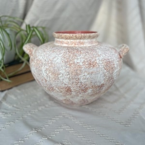 May include: A textured, off-white and terracotta-colored ceramic vase with two small handles. The interior rim is a solid terracotta color. The vase is sitting on a white patterned surface.