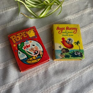 May include: Two vintage comic books: "Popeye" with a red cover and "Bugs Bunny in Double Trouble on Diamond Island" with a yellow cover. The Popeye book features a cartoon of Popeye and a dinosaur. The Bugs Bunny book shows Bugs Bunny in a rocket.