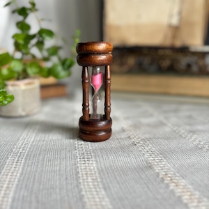 May include: A small hourglass featuring a dark brown wooden frame with a cylindrical top and base connected by four vertical pillars. The central glass chamber contains bright pink sand, which is currently flowing through the narrow neck.