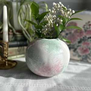 May include: A white ceramic vase with a green and pink speckled glaze. The vase is round and has a wide opening. It is filled with small white flowers.