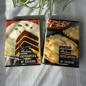 May include: Two vintage cookbooks with yellow spines and colorful covers. The covers feature photos of desserts, including a cake with white frosting and a meringue pie. The titles on the covers read "Meta Given's Modern Encyclopedia of Cooking".