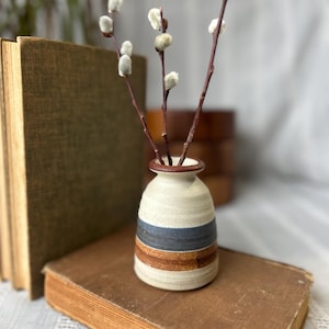 May include: A small, ceramic vase with a cream-colored base, a blue stripe, and a brown stripe. The vase holds several branches with fluffy, white buds. The vase is sitting on a vintage book.